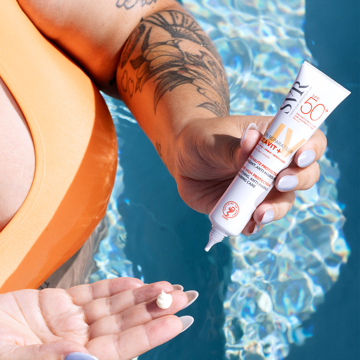 Tubo branco de SVR Cicavit+ Creme SPF50+ sendo aplicado na mão sobre fundo de piscina azul brilhante.
