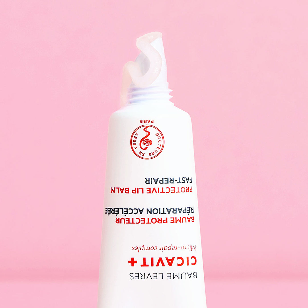 Tubo branco de bálsamo labial Cicavit+ Lèvres com textura transparente saindo da ponta sobre fundo rosa.
