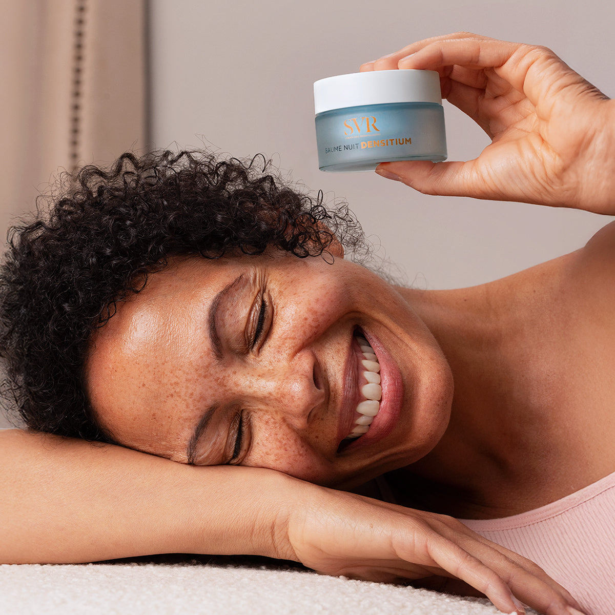 Mulher sorridente segura um frasco azul fosco de SVR Densitium Baume Nuit com tampa branca em fundo neutro.