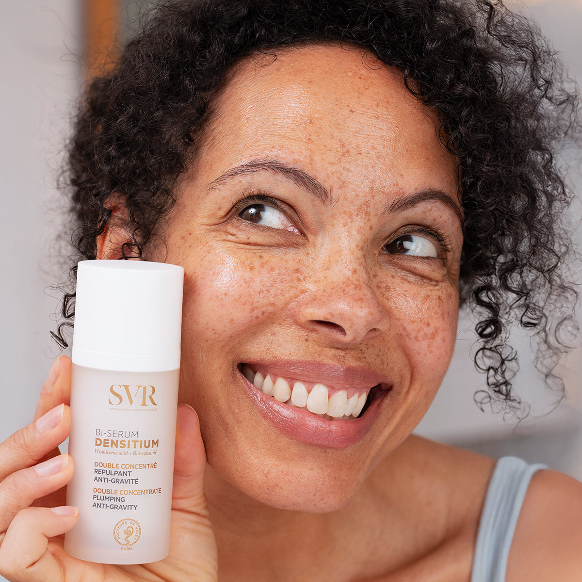 Mulher sorridente segura frasco branco do sérum antienvelhecimento SVR Densitium Bi-Sérum com tampa branca.