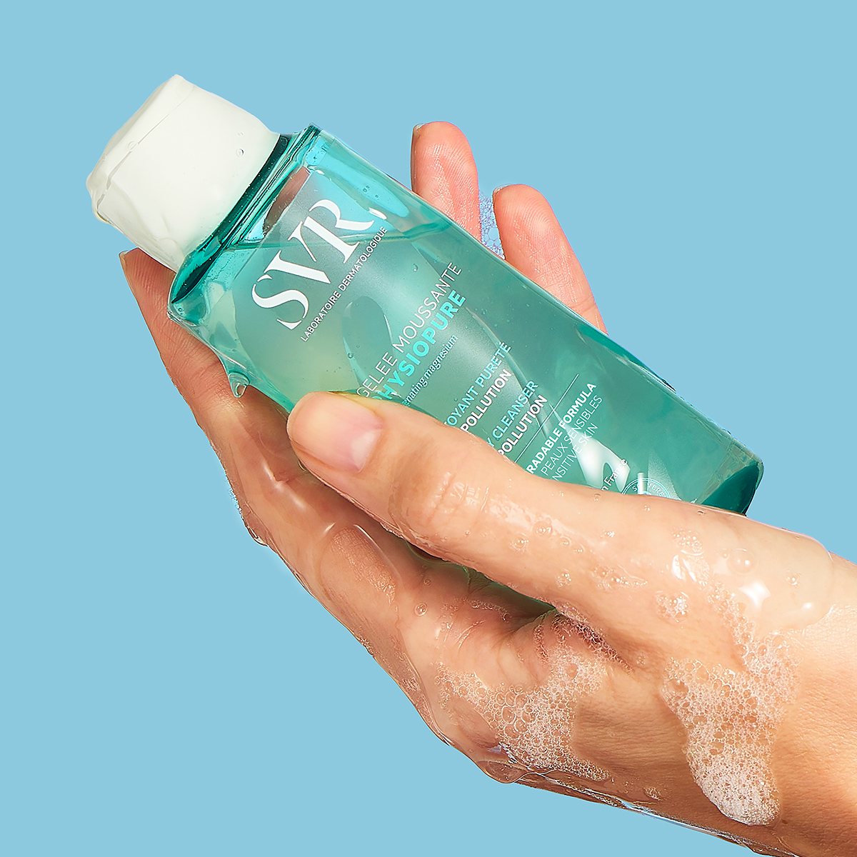 Mão segurando frasco transparente de gel de limpeza SVR PHYSIOPURE azul com tampa branca e espuma suave sobre fundo azul.