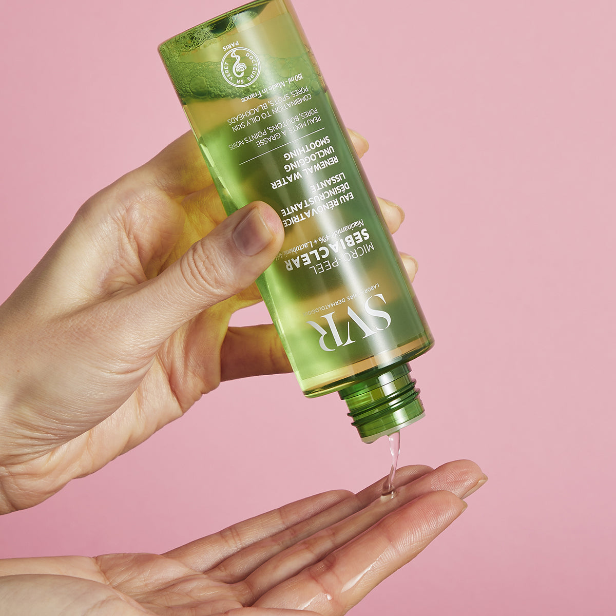Frasco verde transparente de SVR Sebiaclear Micro-Peel sendo aplicado na palma da mão sobre fundo rosa.