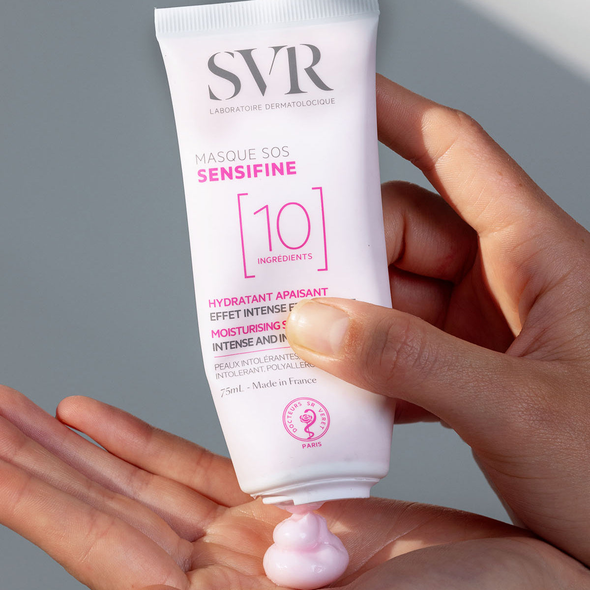Tubo branco de SVR Sensifine Masque SOS com textura cremosa rosada sendo aplicada na palma da mão.