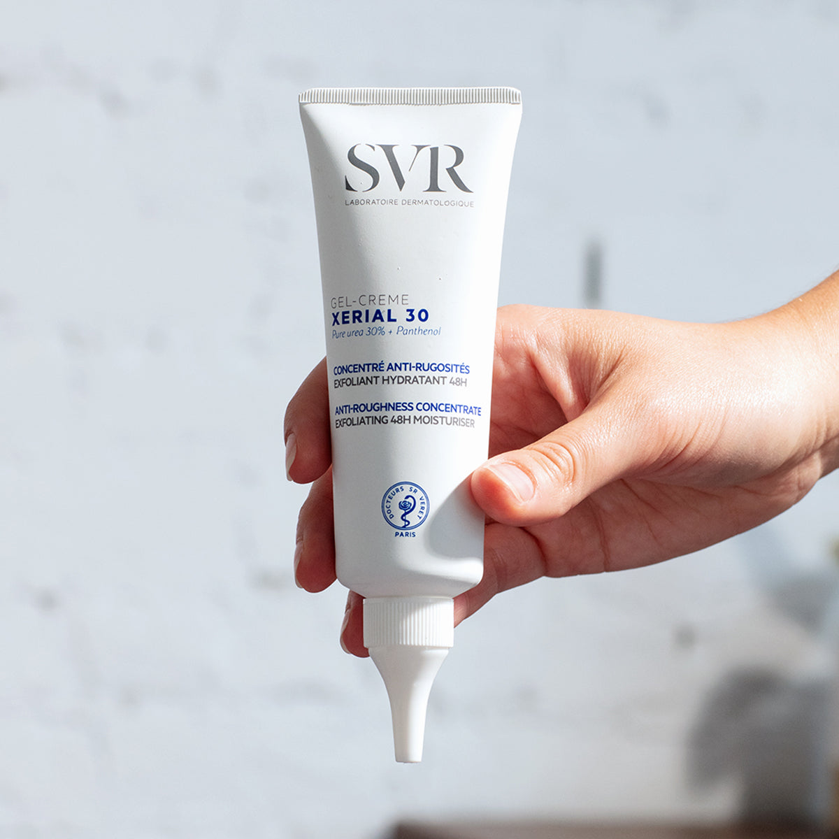 Tubo branco de SVR XERIAL 30 Gel-Crème com aplicador de precisão, segurado por uma mão sobre fundo neutro.