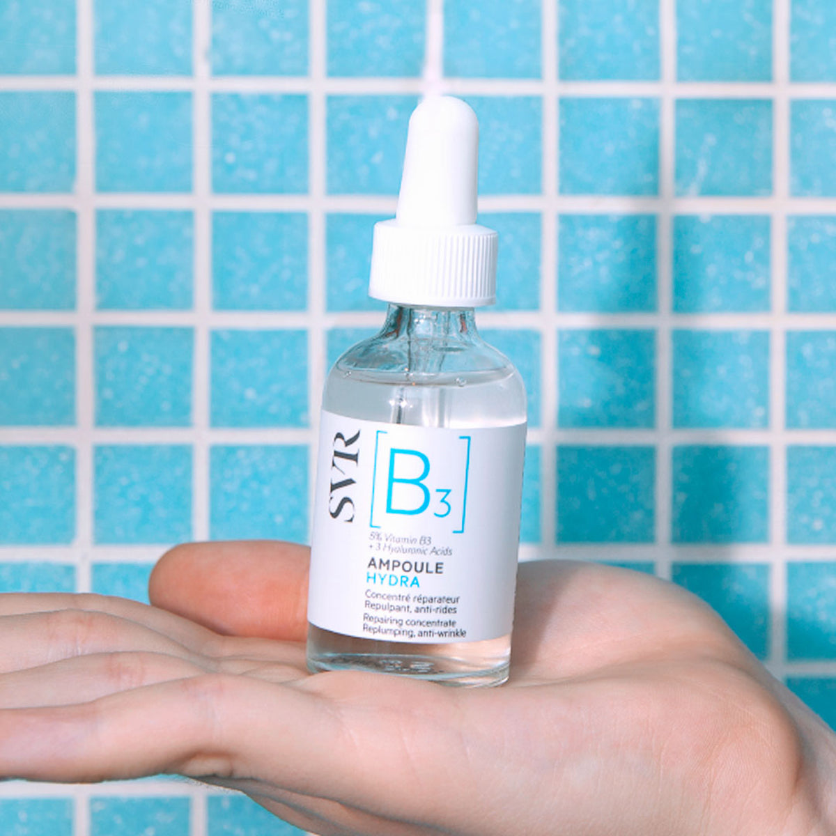 Frasco de vidro transparente SVR Ampoule Hydra com conta-gotas branco, segurado na palma da mão sobre fundo de azulejos azuis.