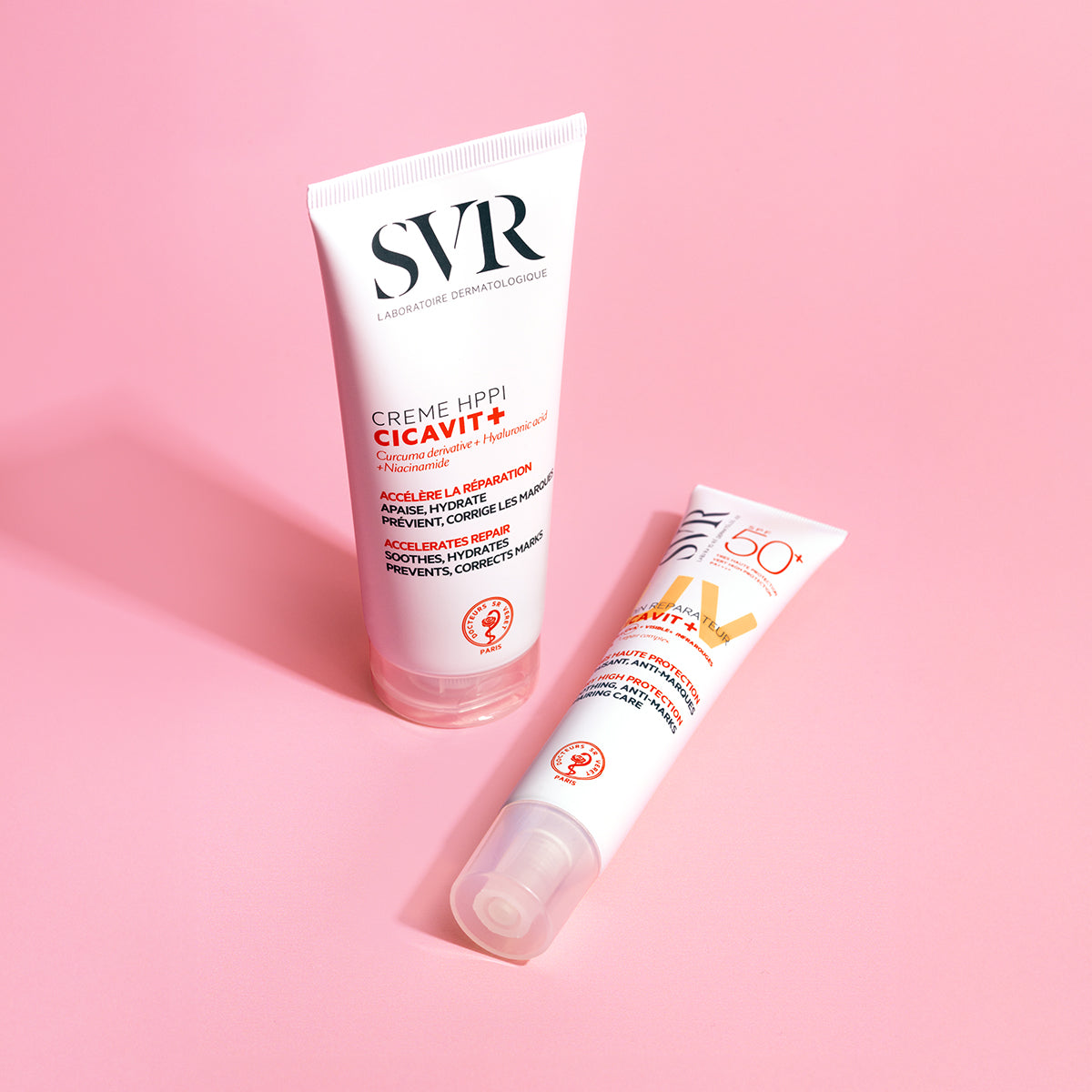 Dois tubos brancos de creme SVR Cicavit+ SPF50+ sobre fundo rosa, um em pé e outro deitado, com detalhes em vermelho e laranja.