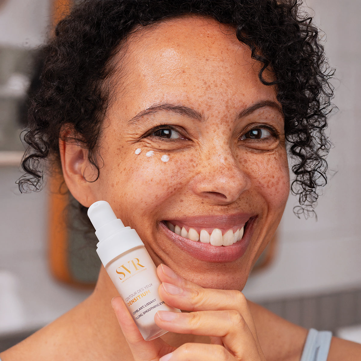Mulher sorridente aplicando creme SVR DENSITIUM Contour des Yeux branco com doseador no contorno dos olhos.