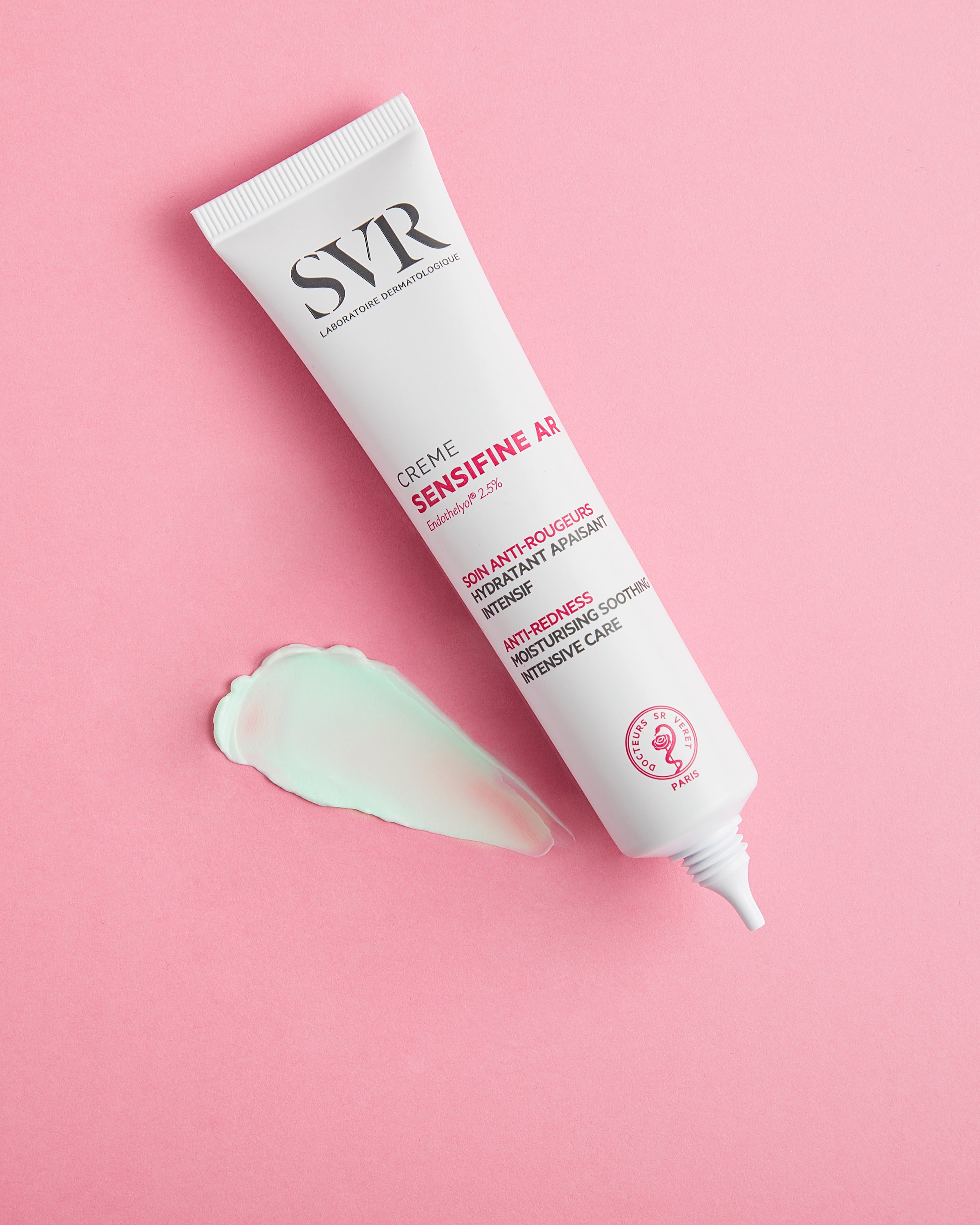 Bisnaga branca de creme SVR Sensifine AR com amostra de textura verde clara sobre fundo rosa.
