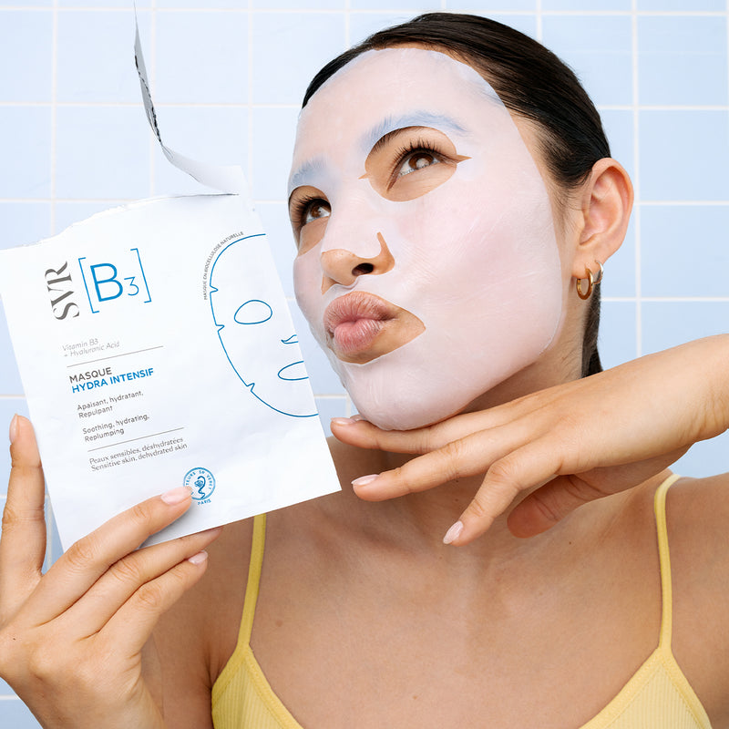 Woman wearing a white SVR B3 hydrating sheet mask, holding the product packaging against a light blue tiled background.