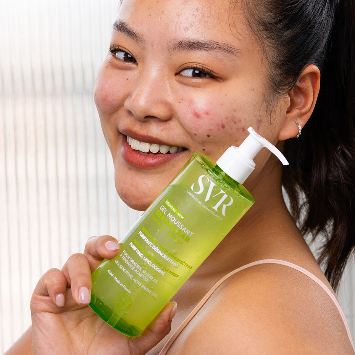 Jovem sorridente segurando frasco de gel de limpeza facial verde SVR Sebiaclear com válvula pump branca.
