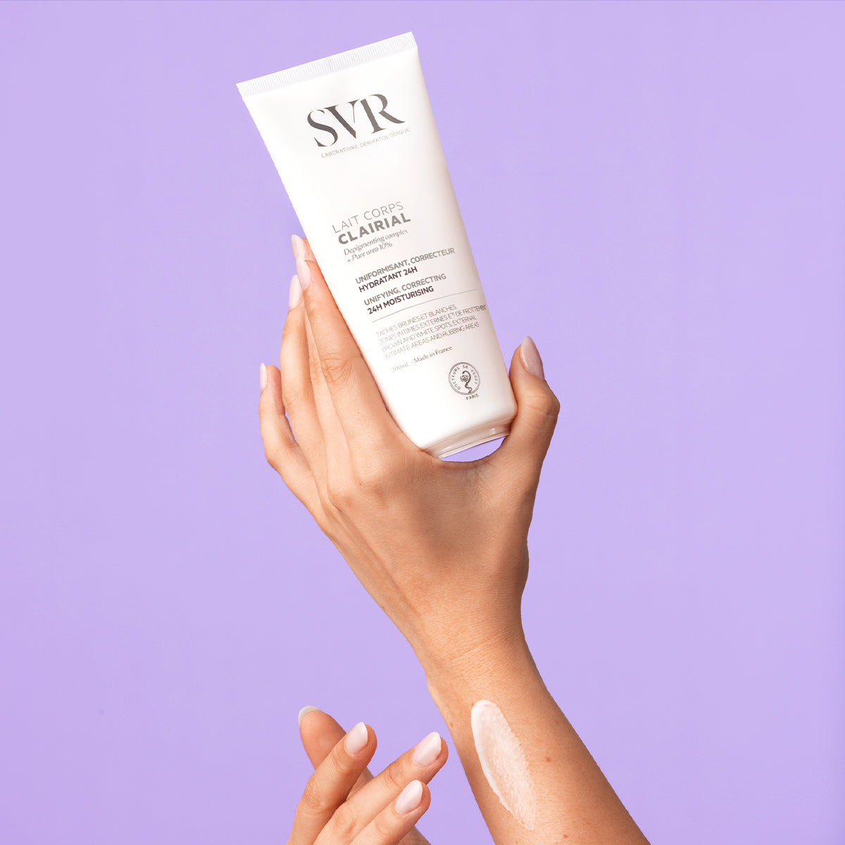 Hand holding a white SVR CLAIRIAL Lait Corps tube with a swatch of moisturizing cream on the forearm, purple background.