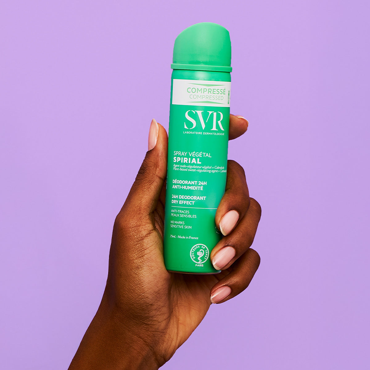 Mão segurando frasco verde de desodorizante SVR Spirial Spray Végétal de 75ml contra fundo lilás.